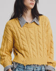 A person in the Denimist Boxy Cable Sweater, a chunky yellow cotton knit, layered over a black and white striped shirt with light blue high-waisted jeans. They have long dark hair and wear black glasses.