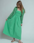 A woman in a Brazeau Tricot Chunky Perforated Cardigan stands against a plain background, holding out one side of her light green dress. She has long light brown hair and wears transparent sandals with colorful decorations.