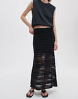 Clad in a sleeveless black top, Rag & Bone’s britt maxi skirt with sheer crochet details, and black sandals, a person stands against a plain light background—an effortlessly summer-ready look.