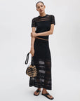 A woman wears the Rag & Bone britt ss crew, a black sheer short-sleeve top, paired with a matching long crochet skirt, black sandals, woven bag, and lightweight cotton sweater against a plain background.