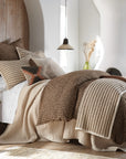 The Amity Home Bruno Duvet Cover King, featuring beige and brown stripes, styled with decorative pillows and a knit throw, brings modern elegance to a bright room with wood accents, a side table, and dried plants in a vase.