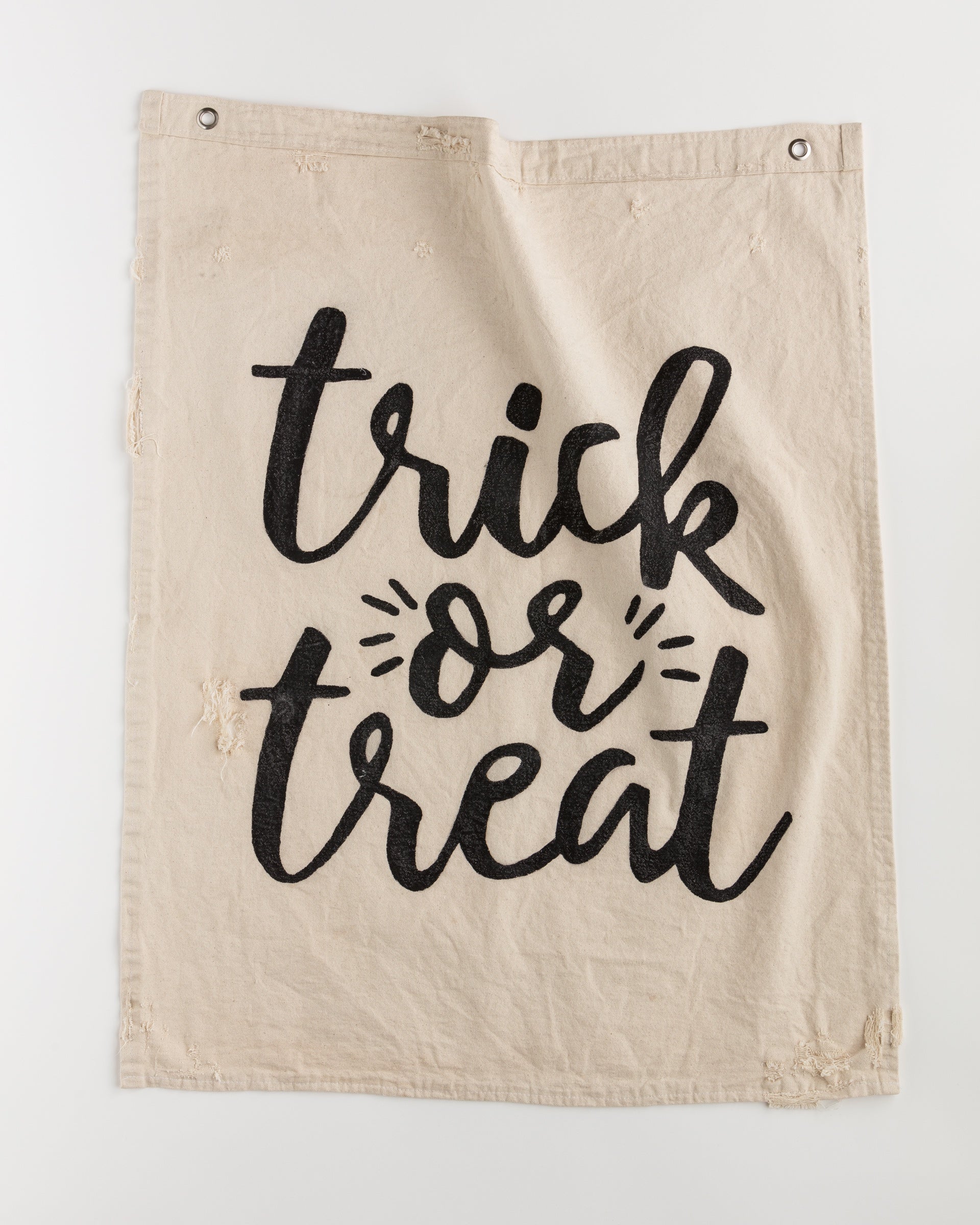 The Tak Design Camp Flags 26x33 in beige fabric showcases the words "trick or treat" in large, bold, black letters. The text is framed by small decorative lines on either side, with frayed edges adding a vintage touch. For easy hanging, two metal grommets are positioned at the top corners, making it an ideal choice for vintage Halloween decor.