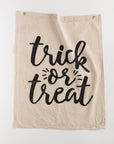 The Tak Design Camp Flags 26x33 in beige fabric showcases the words "trick or treat" in large, bold, black letters. The text is framed by small decorative lines on either side, with frayed edges adding a vintage touch. For easy hanging, two metal grommets are positioned at the top corners, making it an ideal choice for vintage Halloween decor.