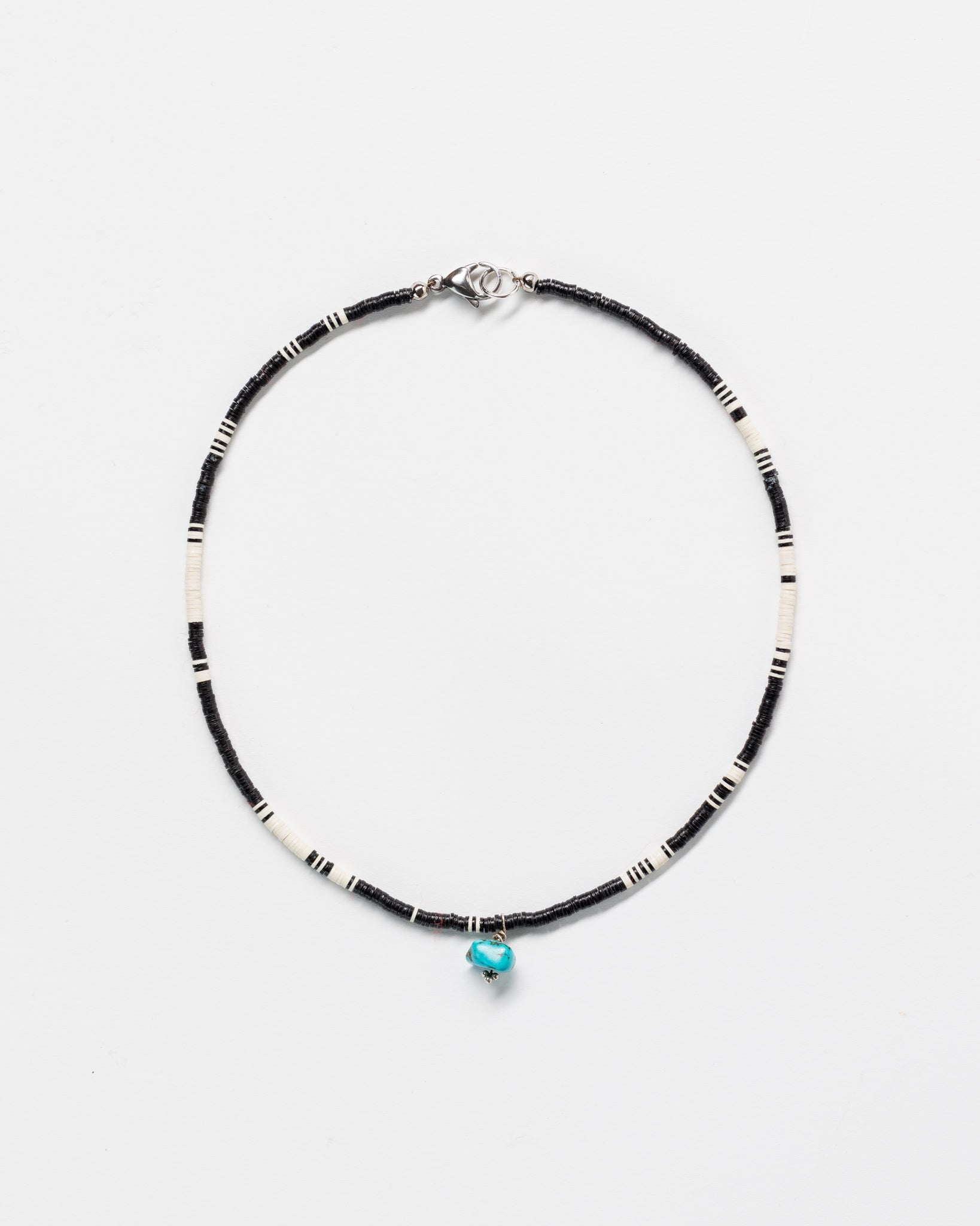 A simple necklace with alternating black and white beads and a single turquoise bead centered below, set against a plain white background, evokes traditional Arizona style.