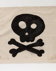 The Camp Flags 26x33 from Tak Design features a distressed beige canvas showcasing a black skull and crossbones design in the center, exuding vintage Halloween charm. The skull's large, round eye sockets and small, round nose add character, while metal grommets in the corners enhance its rustic aesthetic.