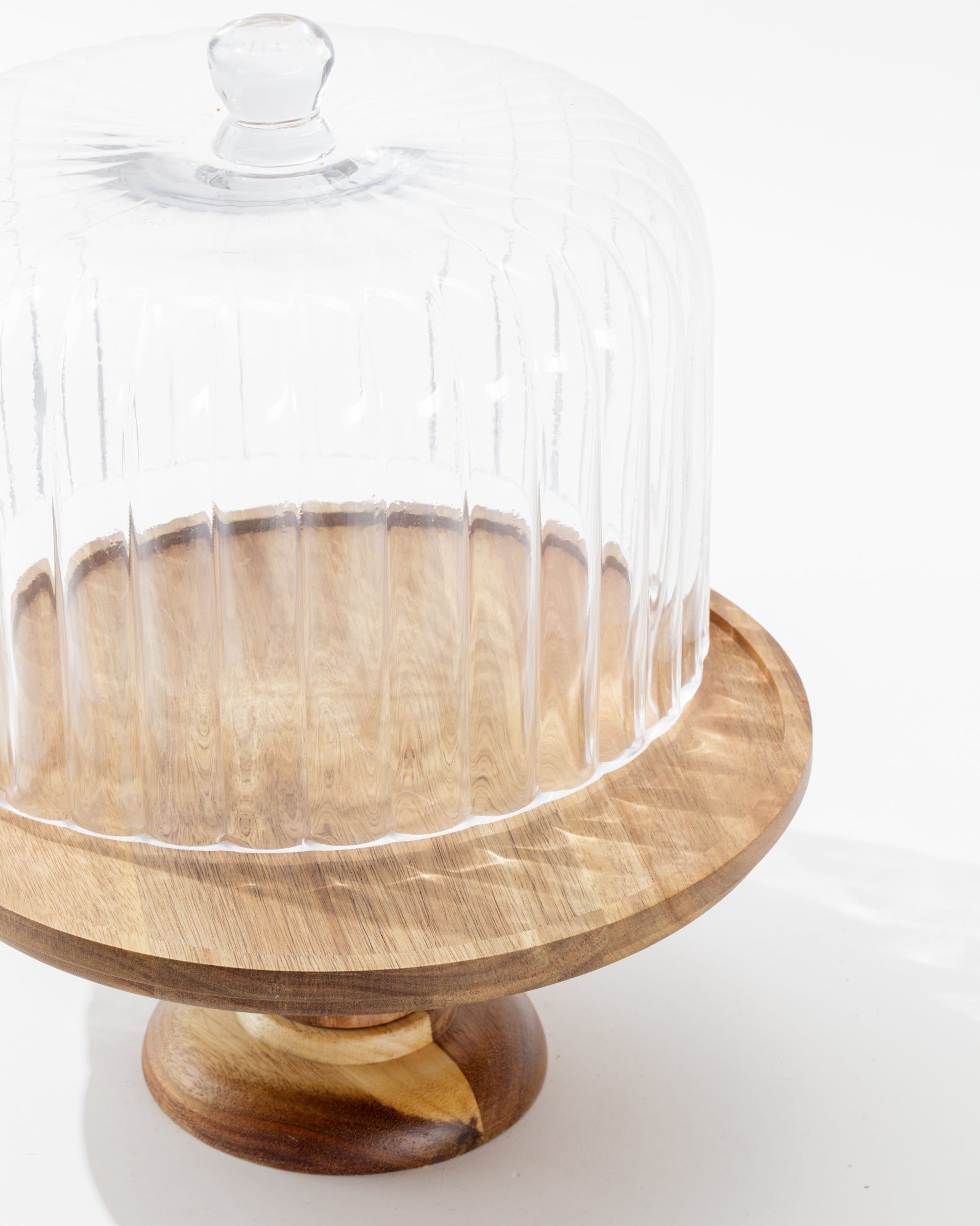 The Creative Co-op Cake Cloche features a wooden pedestal base and a clear, ribbed glass dome. Photographed on a white background, it’s empty and well-lit, highlighting both the rich wood grain and textured glass.