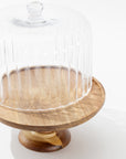 The Creative Co-op Cake Cloche features a wooden pedestal base and a clear, ribbed glass dome. Photographed on a white background, it’s empty and well-lit, highlighting both the rich wood grain and textured glass.