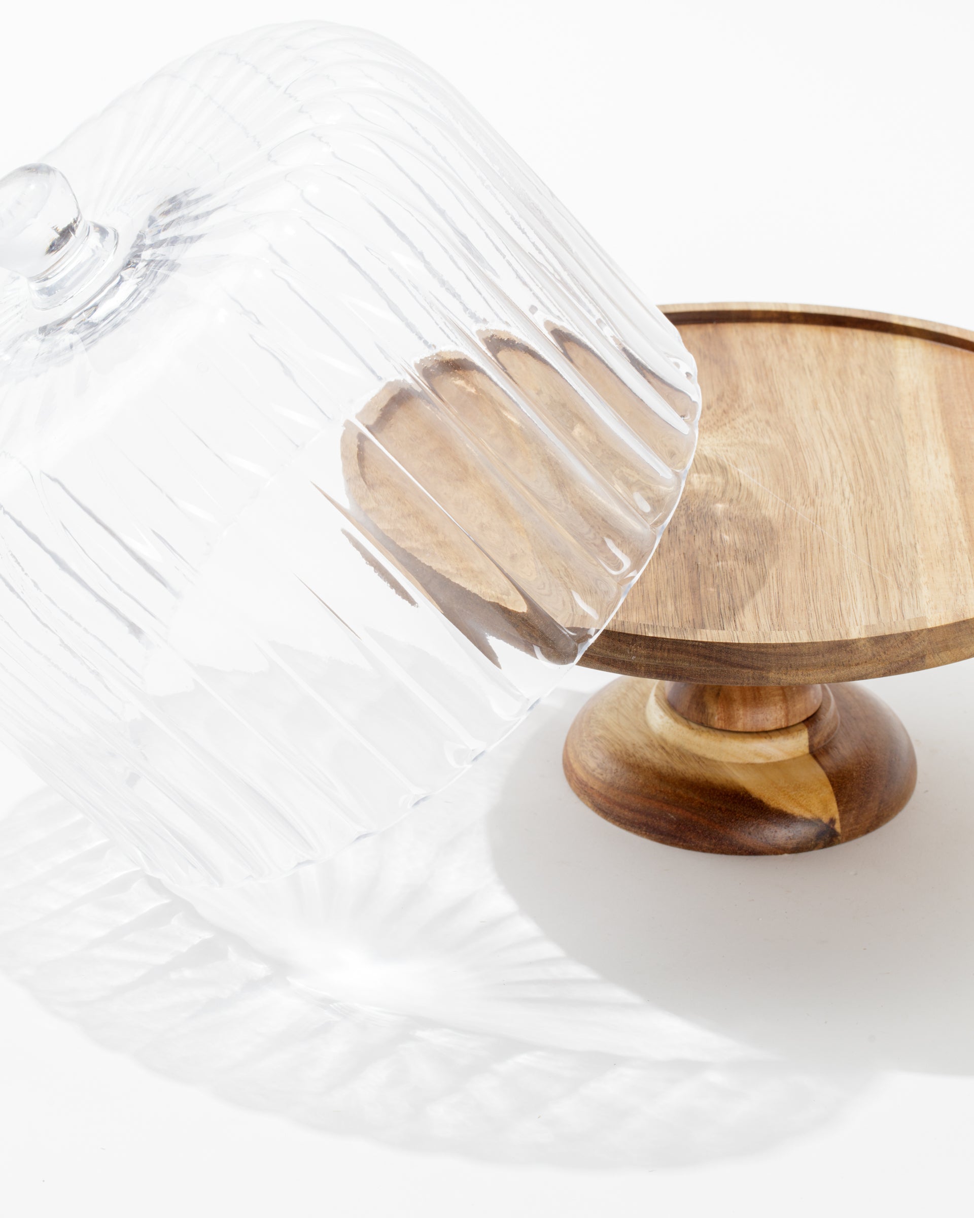 The Creative Co-op Cake Cloche features a wooden pedestal and a tilted glass dome, casting gentle shadows on any surface—an elegant addition to your serveware or collection.