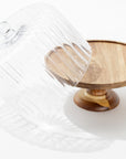 The Creative Co-op Cake Cloche features a wooden pedestal and a tilted glass dome, casting gentle shadows on any surface—an elegant addition to your serveware or collection.