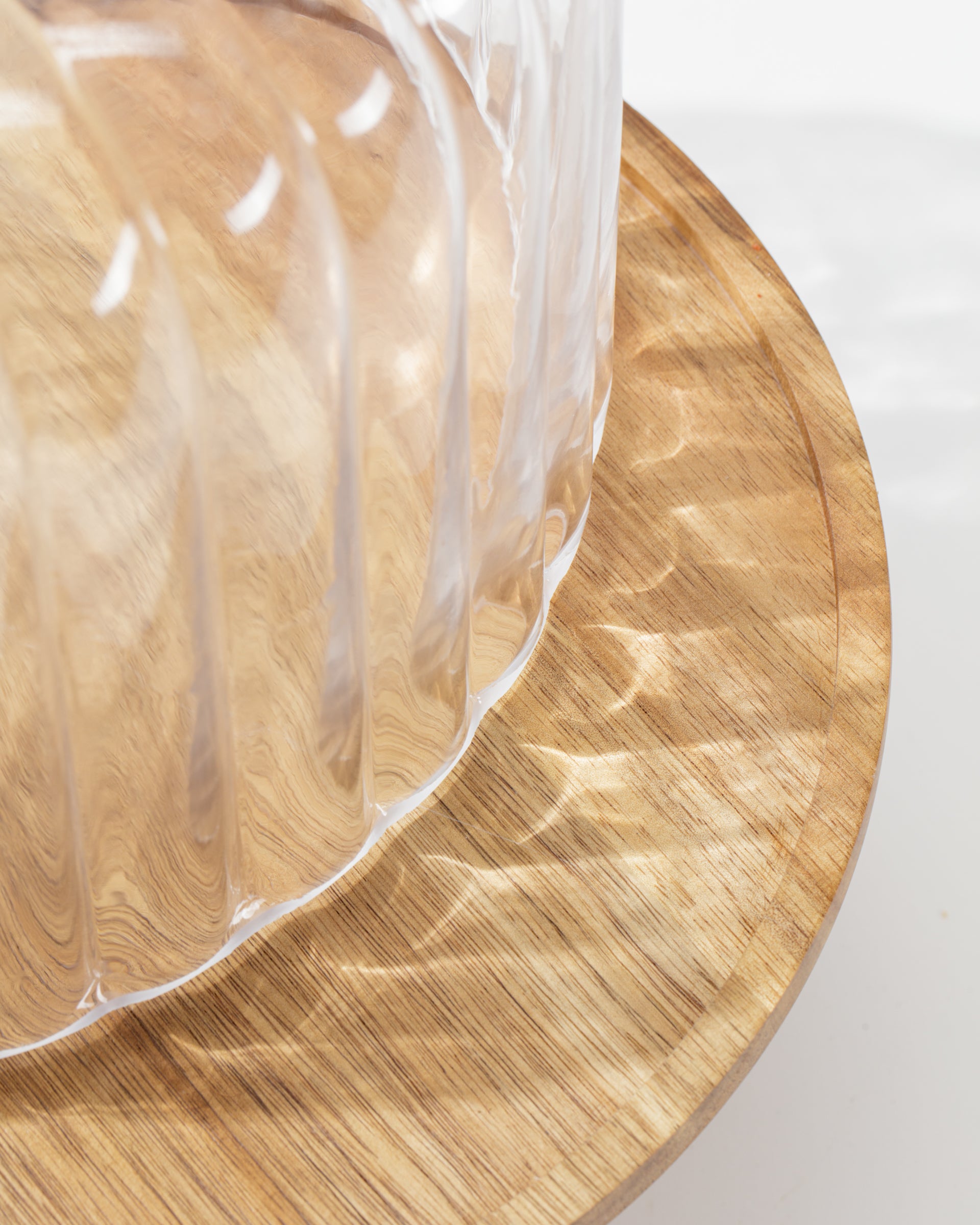 A close-up shows the Creative Co-op Cake Cloche, a clear ribbed plastic cover atop a round wooden stand, showcasing elegant serveware as light casts soft reflections and shadows.