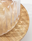 A close-up shows the Creative Co-op Cake Cloche, a clear ribbed plastic cover atop a round wooden stand, showcasing elegant serveware as light casts soft reflections and shadows.