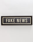 The Street Sign In Black by Spicher And Company features bold white "FAKE NEWS" text and a white border on a rectangular black background, creating eye-catching vintage-inspired wall decor for any room.