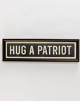 The Street Sign In Black by Spicher And Company features bold "HUG A PATRIOT" lettering on a black and white vintage-style rectangular sign, offering charming wall decor when mounted on a plain white wall.