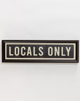 The Street Sign In Black by Spicher And Company features a black rectangular frame with white "LOCALS ONLY" lettering and a thin border, creating striking wall decor when mounted on a white wall.