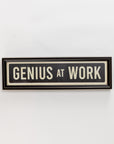 The Spicher And Company Street Sign In Black features "GENIUS AT WORK" in bold capital letters, offering stylish black and white wall decor with a vintage vibe, displayed on a plain white background.