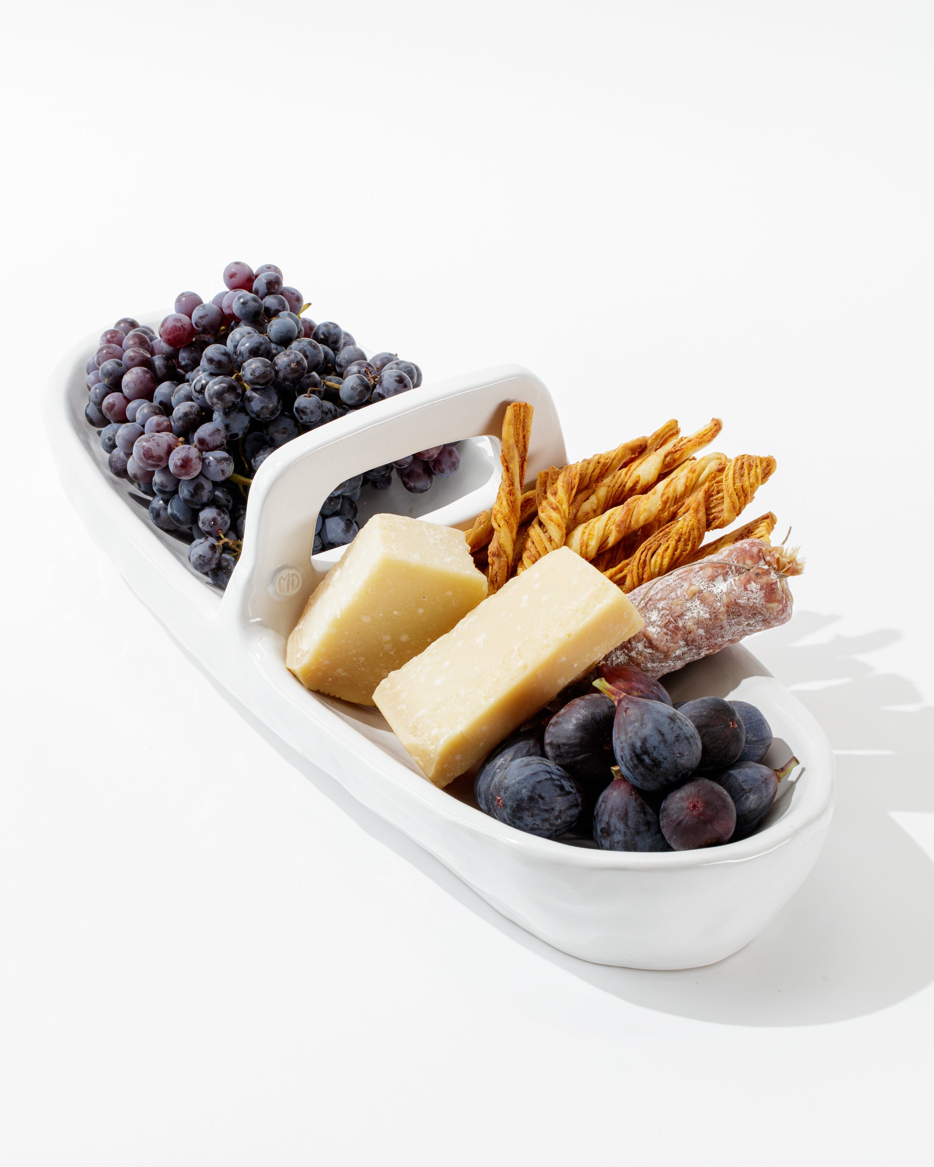 The 4774 Appetizer Platter NO. 774 by Montes Doggett elegantly holds a selection of grapes, two large pieces of cheese, breadsticks, and a piece of cured sausage. The food is artistically arranged against a white background, highlighting the platter's traditional craftsmanship and artisanal charm.