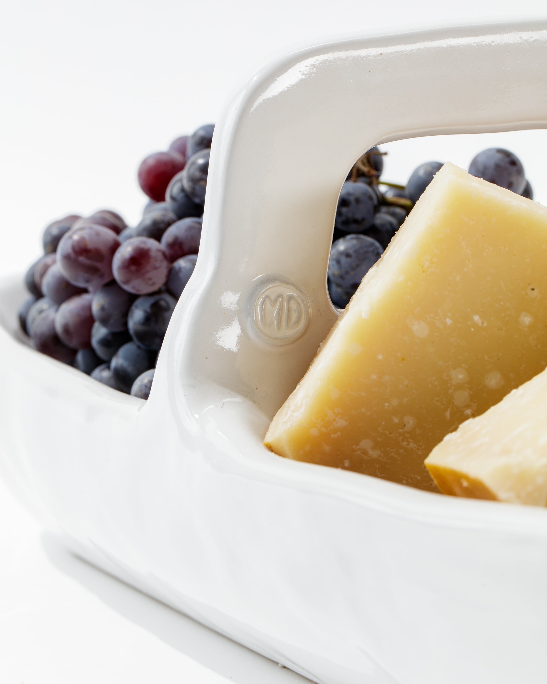 A close-up image of a white *handmade ceramic* basket from the 4774 Appetizer Platter NO. 774 collection by Montes Doggett, containing chunks of hard cheese and a bunch of grapes. The basket handle has the initials "MD" embossed on it, showcasing traditional techniques. The background is plain white.