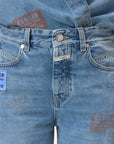 Close-up of a person wearing CLOSED's Nikka Denim jeans and matching shirt in light blue salt-and-pepper wash, featuring colorful "CLOSED" text patches and crafted with eco-conscious materials.