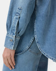 A close-up shows a person wearing the CLOSED Oversized Shirt in mid-blue denim with a curved hem, buttoned cuff, jeans, and embroidered patches. Their hand rests relaxed by their side.