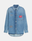 The CLOSED Oversized Shirt is a light blue denim piece featuring a pointed collar, front pocket, and multicolored printed and embroidered patches with text designs on the chest and pocket areas.