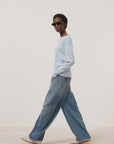 A person in dark sunglasses, wide-leg blue jeans, white sneakers, and the CLOSED Basic Long Sleeve Tee with light blue and white stripes walks against a plain light background.