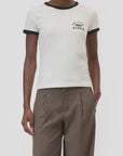 A person wears the CLOSED Slim T-Shirt—white with black collar and sleeve cuffs, featuring a small "Closed" logo on the chest—paired with brown pleated trousers against a plain background.