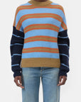 A person models the CLOSED Crewneck LS Knit, a colorful striped wool sweater in blue, red, mustard, and navy, paired with light blue jeans against a plain white background.
