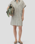 A person in the CLOSED Turn-up Shirtdress with short sleeves, stripes, and mother-of-pearl buttons, paired with flip-flops and holding a green and beige woven bag draped with scarves, stands against a plain light background.