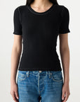 A person wears the AMO Denim Cherish Tee, a fitted black short sleeve ribbed cotton top, with blue jeans against a plain light background. Their face is partially out of frame.