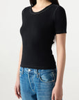 A woman wears the Cherish Tee by AMO Denim, a fitted black ribbed cotton short sleeve top, paired with blue jeans. She is shown waist up against a light background with long straight hair and her face partially out of frame.