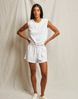 A woman poses against a plain backdrop wearing the Perfectwhitetee Carli Muscle Tank, white drawstring shorts, and white sneakers. She stands hands on hips, looking toward the camera.