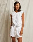 A woman with long dark hair stands against a textured backdrop, wearing the Perfectwhitetee Carli Muscle Tank in white terry cloth and matching drawstring shorts. She poses with a neutral expression, one arm relaxed at her side.