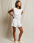 A woman stands before a neutral fabric backdrop, wearing the Perfectwhitetee Carli Muscle Tank and matching drawstring shorts in white, paired with white sneakers. She looks to the side with her hands behind her back.