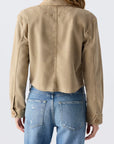 A person with long, light brown hair is seen from the back wearing the AMO Denim Carly Jacket in tan, paired with blue jeans featuring distressed pocket details.