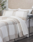 A neatly made bed features the Amity Home Chalmers Quilt Nat K in beige and white buffalo check, with two large pillows and a light wood headboard. A nightstand holds books, a decorative item, and a potted plant sits beside the bed.