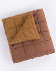 The Spencer Quilt by Amity Home is a reversible, folded, quilted blanket with a grid pattern in brown and mustard yellow slub cotton. Visible square stitching stands out, and the quilt is displayed against a white background.