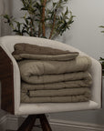 A modern upholstered chair displays three folded Amity Home Spencer Quilts in beige and brown, with green leafy plants set against a light gray wall.