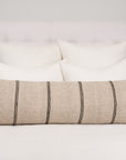 A neatly made bed with a white tufted headboard, four white pillows, and the Amity Home Rhodes Body Pillow Sham—a natural linen bolster with black stripes—sits against a light grey wall. Bedside tables hold books and decor on either side.