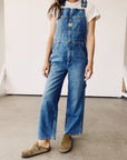 A person stands against a plain background wearing The Miner Overalls by Hey Gang, featuring a unisex fit in vintage washed denim, paired with a white T-shirt. They have on brown casual shoes, with their hands by their sides.