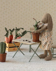 A Maileg bunny in a floral dress stands by a small table, potting the Maileg Flower Pot. Nearby are a wooden crate with fabric carrots, two chairs, and a potted fabric cactus on the floor.