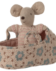 The Maileg Baby Mouse in Carry Cot by Maileg features a baby mouse in a knitted suit, snug in a patterned cot with blue and brown flowers, clutching the basket’s handle.
