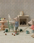 Two Maileg toy mice in striped shirts tidy a cozy room: one vacuums pebbles, the other lifts a Vintage Chair. The dollhouse-like space is filled with charming mice furniture, patterned decor, dishes on a table, and a glowing fireplace.
