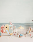 Maileg's Little Brother and friends don beach attire to relax and play by colorful cabanas. A stuffed cat perches nearby, all set for imaginative adventures with the Maileg Little Brother /Beach Cabana set.