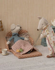 Maileg Grandma & Grandpa Mice by Maileg relax in a cozy room—Grandma naps in a matchbox bed, Grandpa vacuums in his robe. Nearby sit a lamp, tea set, and teapot on a blue cabinet.
