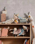 A wooden shelf shows Maileg plush toys and furniture, featuring the Maileg Medium Mouse in a nightgown as a cozy bedtime friend, with small toy beds, miniature furniture, books, and a tall stuffed giraffe against a plain light gray wall.