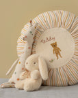 A Maileg Plush Bunny Mini by Maileg sits beside a round, striped cushion featuring an embroidered teddy bear and "Teddy," plus a fabric giraffe with colorful polka dots—an adorable gift for kids who love soft stuffed animals.