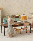 Two Maileg mice in dresses stand in a vintage kitchen, with one serving cake on the Maileg Long Dining Table set with plates and a teapot. Dollhouse furniture and a retro fridge finish the charming scene.
