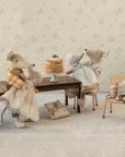 Five Maileg stuffed toy mice enjoy a stack of pancakes and a gift around the Maileg Long Dining Table. Two sit at the table, one cooks at a play stove, and two stand nearby, all in cute, detailed outfits. Brand: Maileg.