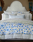 A neatly made bed features John Robshaw's Cala Duvet, a blockprint bouquet headboard, blue and white floral bedding, decorative pillows, and wooden nightstands with lamps and flowers for a cozy, garden-inspired room in soothing greens and blues.