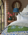 A cozy bedroom features a John Robshaw Cala Duvet in greens and blues, a green floral blanket, a side table with a vase of pink blockprint bouquet flowers, a blue lamp, decorative mirror, and an arched window with patterned curtains.
